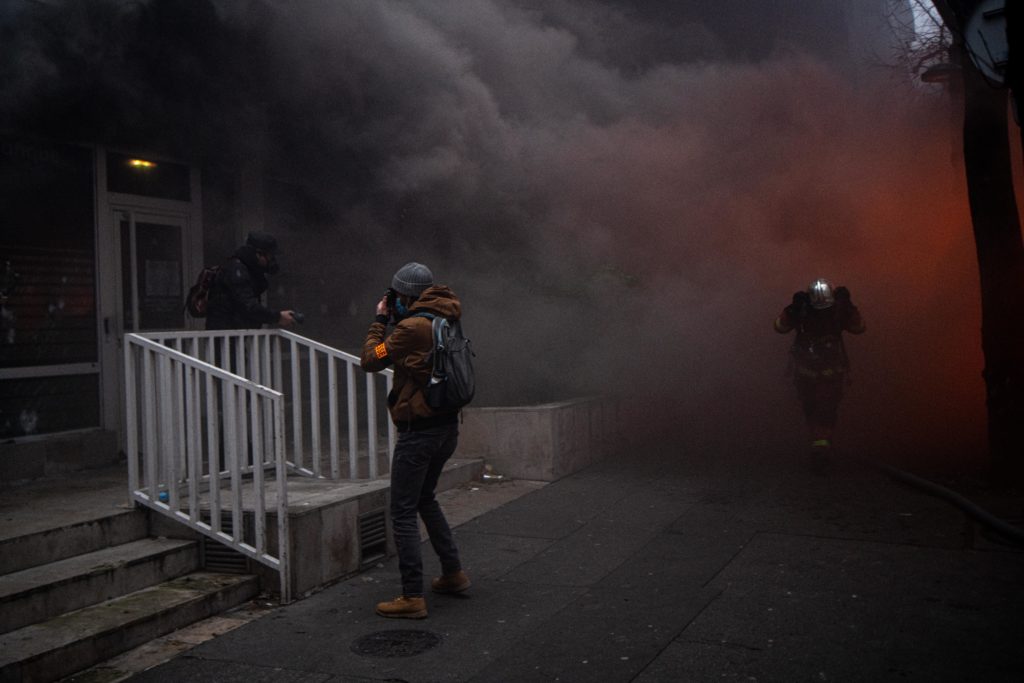 Un photographe en plein travail face à un incendie d'une agence du crédit mutuel avenue Gambetta à Paris, tandis qu'un pompier traverse la fumée pour aller combattre l'incendie. lors de la manifestation du 5 décembre contre le projet de loi dit Sécurité globale.