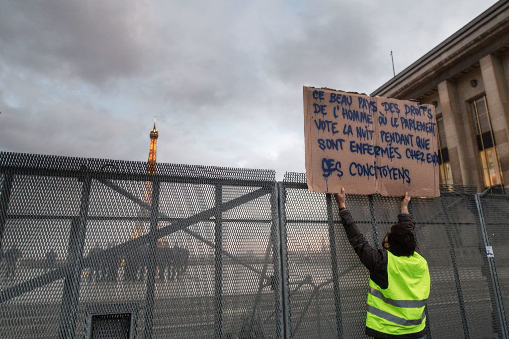 Le 21 novembre 2020, un homme brandit une pancarte mettant en cause l'heure choisie pour le vote de l'article 24 de la proposition de loi Sécurité globale face aux CRS sur le parvis des droits de l'homme derrière une barrière en métal.