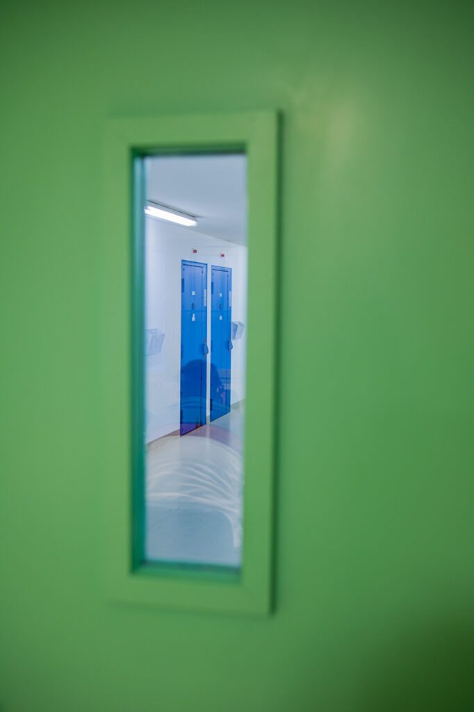 Un patient en cours d'entretien avec l'EMOT est visible dans le reflet d'une fenêtre avec en fond deux portes de cellules.